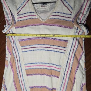 May include: Cream-colored tunic with a V-neck and horizontal stripes in tan, pink, and blue. The tunic has a loose fit and is approximately 18 inches wide. The fabric appears to be a lightweight woven material, suitable for warm weather.