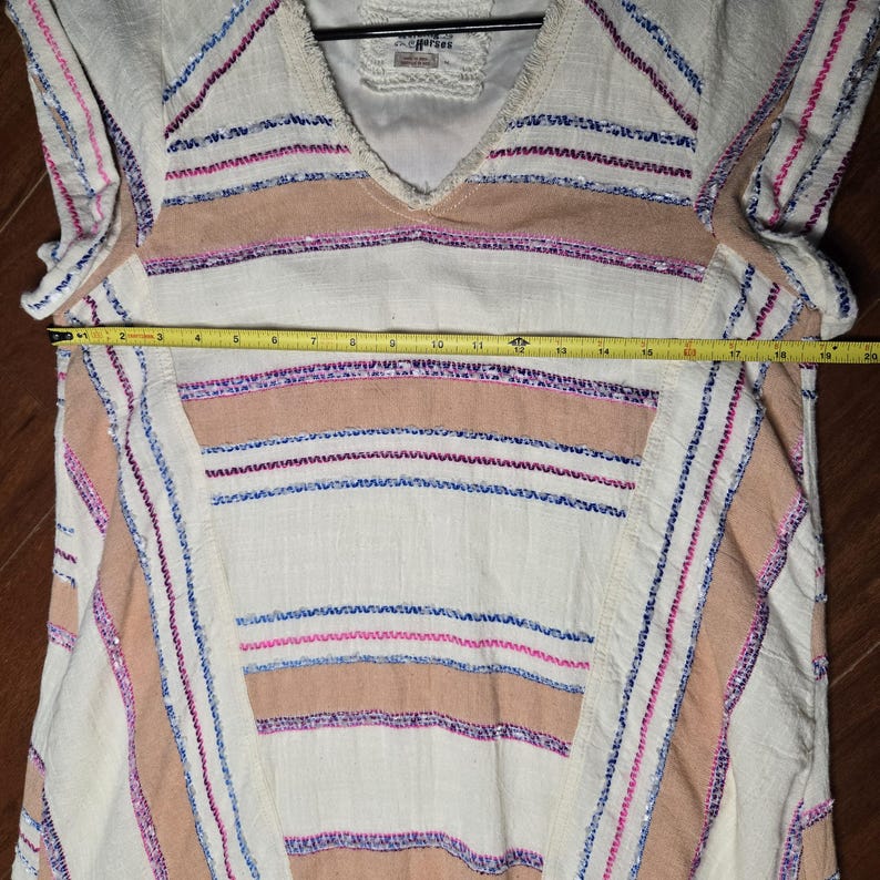 May include: Cream-colored tunic with a V-neck and horizontal stripes in tan, pink, and blue. The tunic has a loose fit and is approximately 18 inches wide. The fabric appears to be a lightweight woven material, suitable for warm weather.