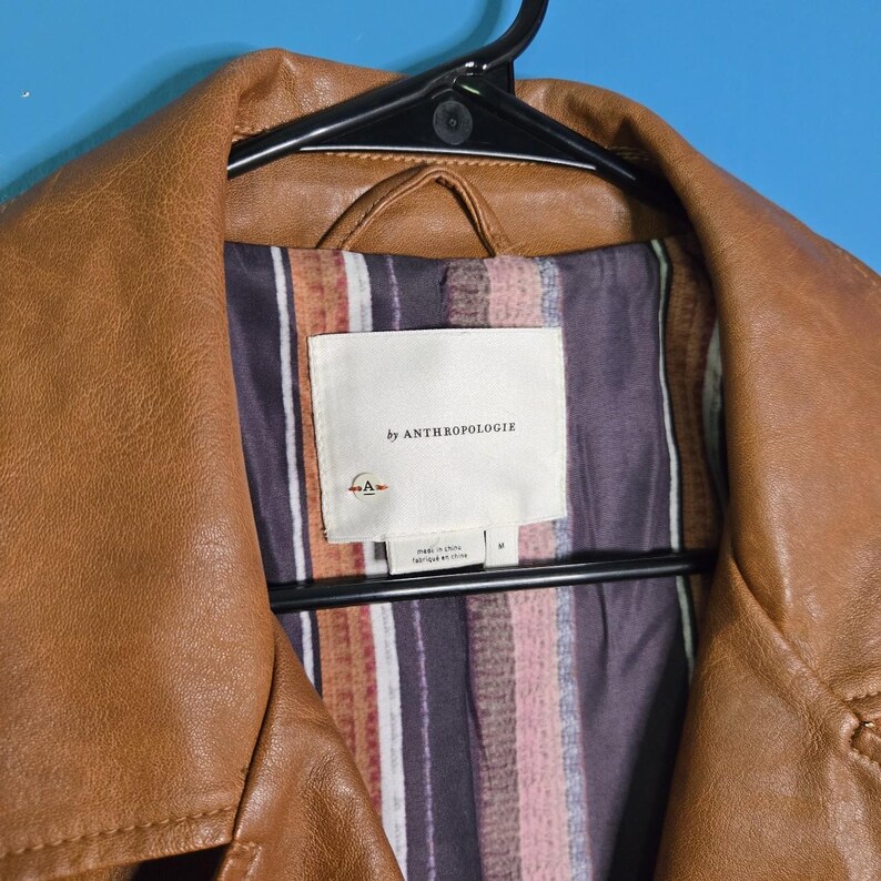 May include: A brown leather jacket hanging on a black hanger. The jacket has a collar and a label that reads "by ANTHROPOLOGIE". The lining is patterned with vertical stripes in shades of brown, pink, and purple.