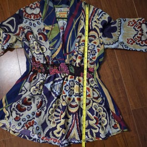 Pode incluir: Uma t&uacute;nica da marca Johnny Was com uma estampa floral e paisley multicolorida em tons de azul, vermelho e creme. A pe&ccedil;a tem decote em V, mangas tr&ecirc;s quartos e cintura el&aacute;stica franzida. Uma fita m&eacute;trica amarela indica 66 cm de comprimento.