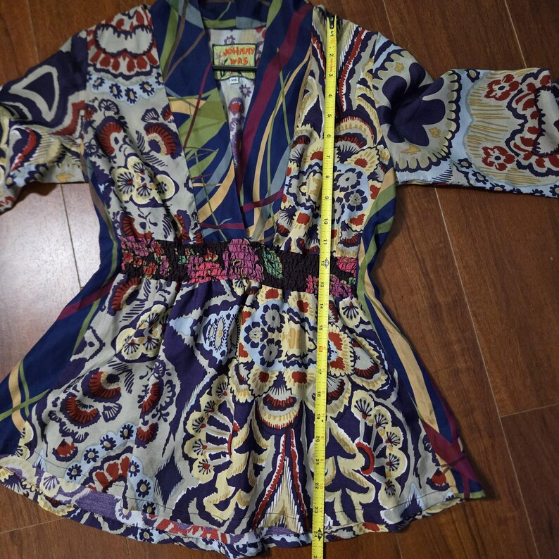Pode incluir: Uma t&uacute;nica da marca Johnny Was com uma estampa floral e paisley multicolorida em tons de azul, vermelho e creme. A pe&ccedil;a tem decote em V, mangas tr&ecirc;s quartos e cintura el&aacute;stica franzida. Uma fita m&eacute;trica amarela indica 66 cm de comprimento.