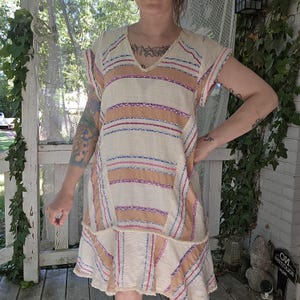 May include: Cream-colored, striped dress with a V-neck and short sleeves. The dress features horizontal stripes in shades of tan, pink, and blue. The hemline has a ruffled detail. The fabric appears to be a lightweight, textured material.