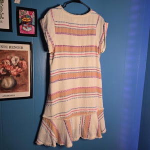 May include: A cream-colored dress with horizontal stripes in shades of tan, pink, and blue. The dress has short sleeves and a ruffled hem. The fabric appears to have a textured, woven quality. The dress is hanging on a black hanger.