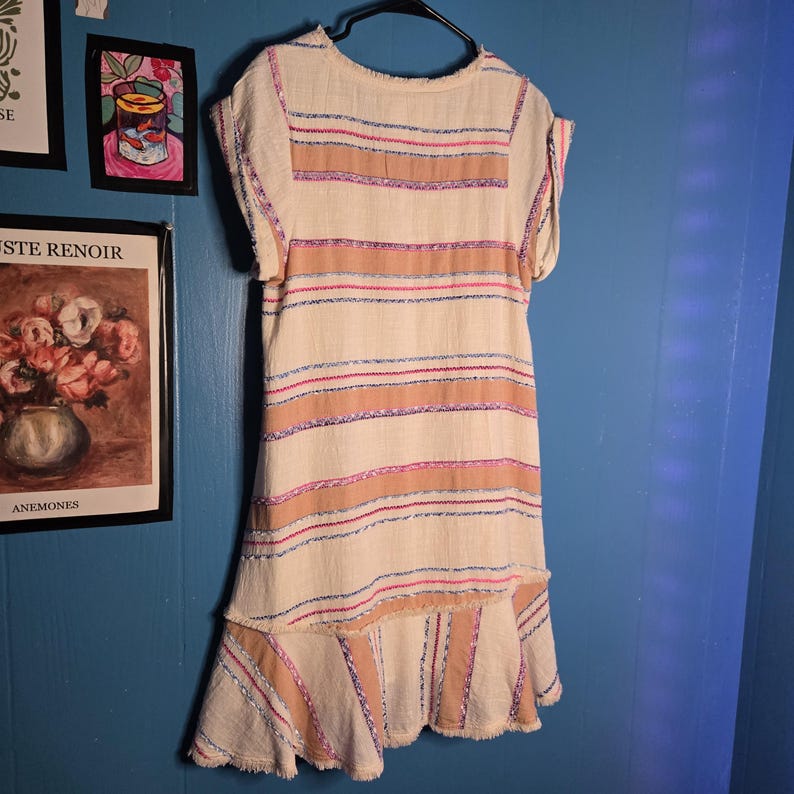 May include: A cream-colored dress with horizontal stripes in shades of tan, pink, and blue. The dress has short sleeves and a ruffled hem. The fabric appears to have a textured, woven quality. The dress is hanging on a black hanger.