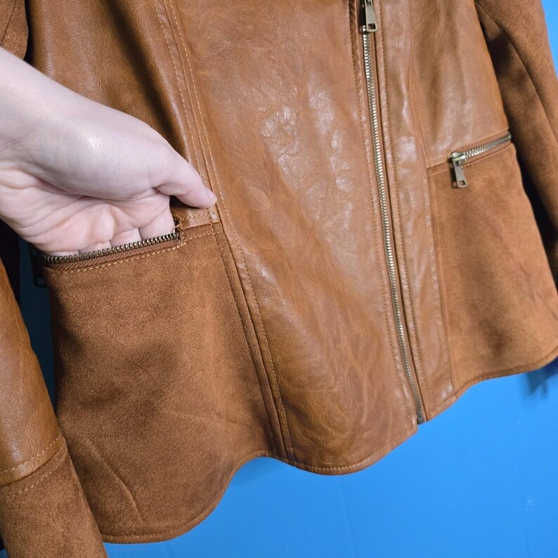 May include: A brown leather jacket with a suede-like texture on the pockets and a full-length zipper. The jacket has a classic design with a tailored fit and a small zippered pocket on the chest.