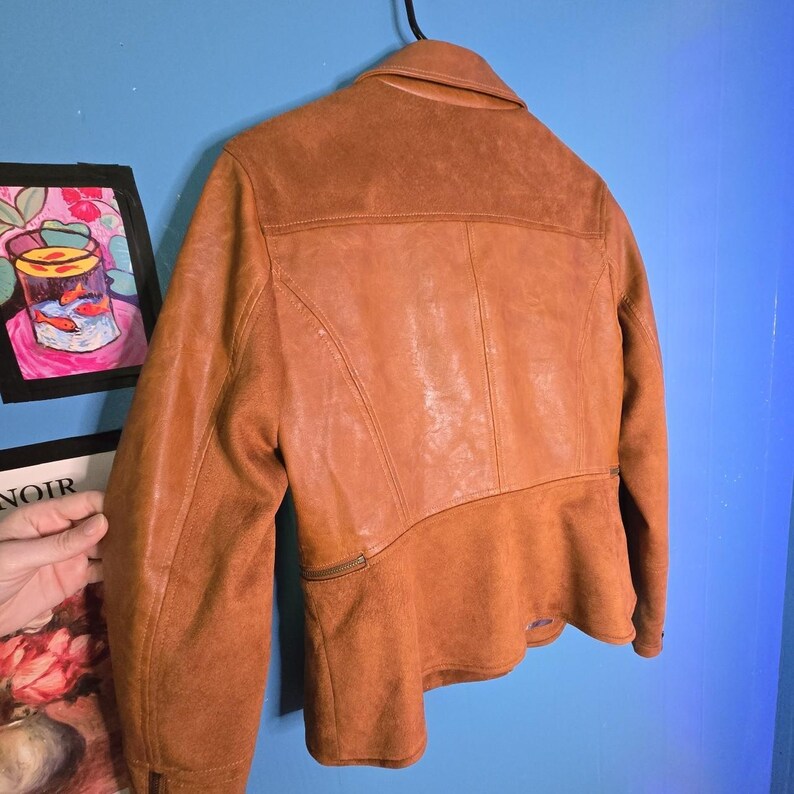 May include: A brown leather jacket with a suede-like texture on the lower back and sleeves. The jacket has a collar, a zipper, and a panel design on the back. The jacket is hanging on a black hook against a blue wall.