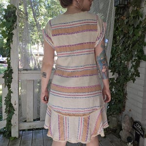 May include: A beige dress with horizontal stripes in shades of brown, pink, and blue. The dress has a tiered skirt and short sleeves. The fabric appears to be a lightweight, textured material, with a fringed hemline.
