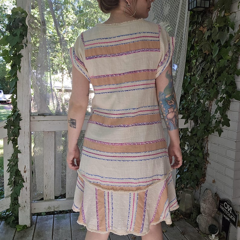 May include: A beige dress with horizontal stripes in shades of brown, pink, and blue. The dress has a tiered skirt and short sleeves. The fabric appears to be a lightweight, textured material, with a fringed hemline.