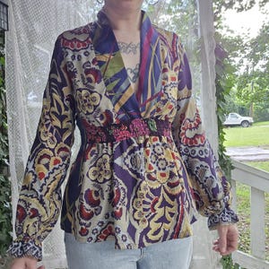 Pode incluir: Uma blusa de manga comprida com decote em V profundo e cintura el&aacute;stica franzida. A pe&ccedil;a &eacute; decorada com um vibrante padr&atilde;o floral e paisley multicolorido em tons de roxo, vermelho, amarelo e creme, com mangas compridas e largas.