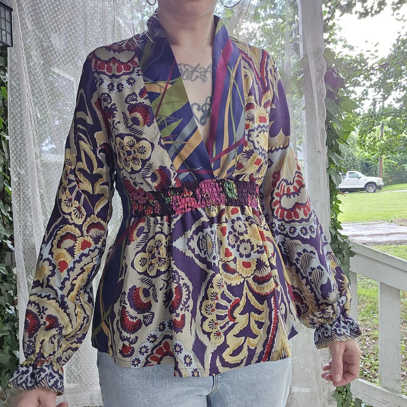 Pode incluir: Uma blusa de manga comprida com decote em V profundo e cintura el&aacute;stica franzida. A pe&ccedil;a &eacute; decorada com um vibrante padr&atilde;o floral e paisley multicolorido em tons de roxo, vermelho, amarelo e creme, com mangas compridas e largas.
