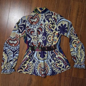 Pode incluir: Uma blusa de manga comprida com um padr&atilde;o floral e paisley multicolorido vibrante em tons de roxo escuro, azul claro, amarelo e vermelho. A pe&ccedil;a inclui um decote alto, uma cintura el&aacute;stica franzida e punhos de manga com folhos.