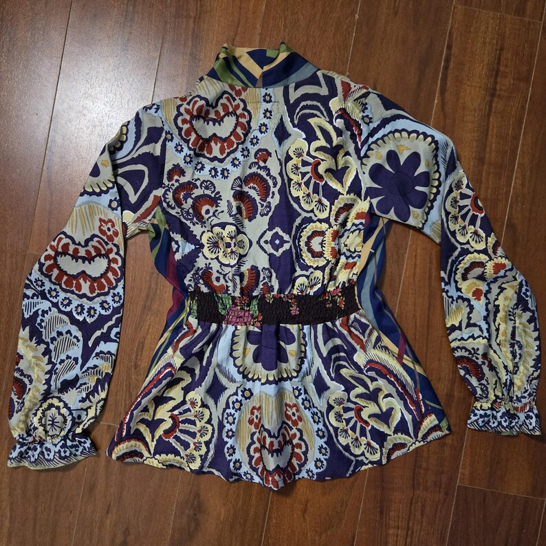 Pode incluir: Uma blusa de manga comprida com um padr&atilde;o floral e paisley multicolorido vibrante em tons de roxo escuro, azul claro, amarelo e vermelho. A pe&ccedil;a inclui um decote alto, uma cintura el&aacute;stica franzida e punhos de manga com folhos.