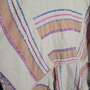 May include: Close-up of a cream-colored fabric with horizontal stripes in shades of tan, pink, and blue. The fabric has a textured appearance with visible stitching and fringed edges, suggesting a woven textile.