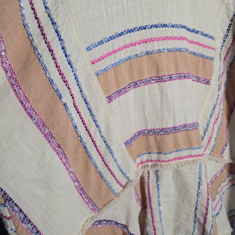 May include: Close-up of a cream-colored fabric with horizontal stripes in shades of tan, pink, and blue. The fabric has a textured appearance with visible stitching and fringed edges, suggesting a woven textile.