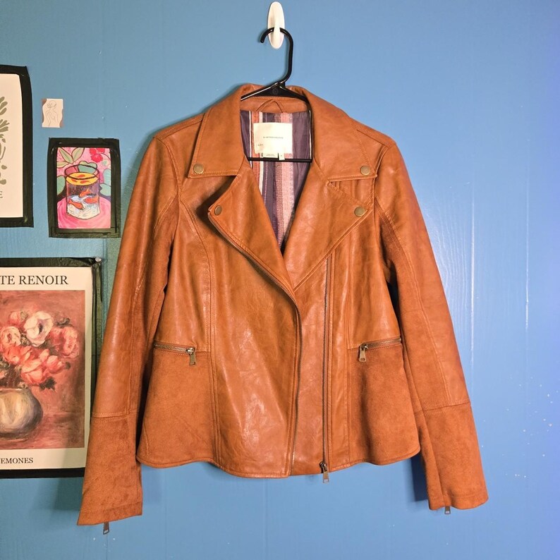 May include: A brown leather jacket with a classic biker style. The jacket features a notched collar, asymmetrical zipper closure, and multiple zippered pockets. The interior lining has a striped pattern. The jacket is hanging on a black hanger against a blue wall.