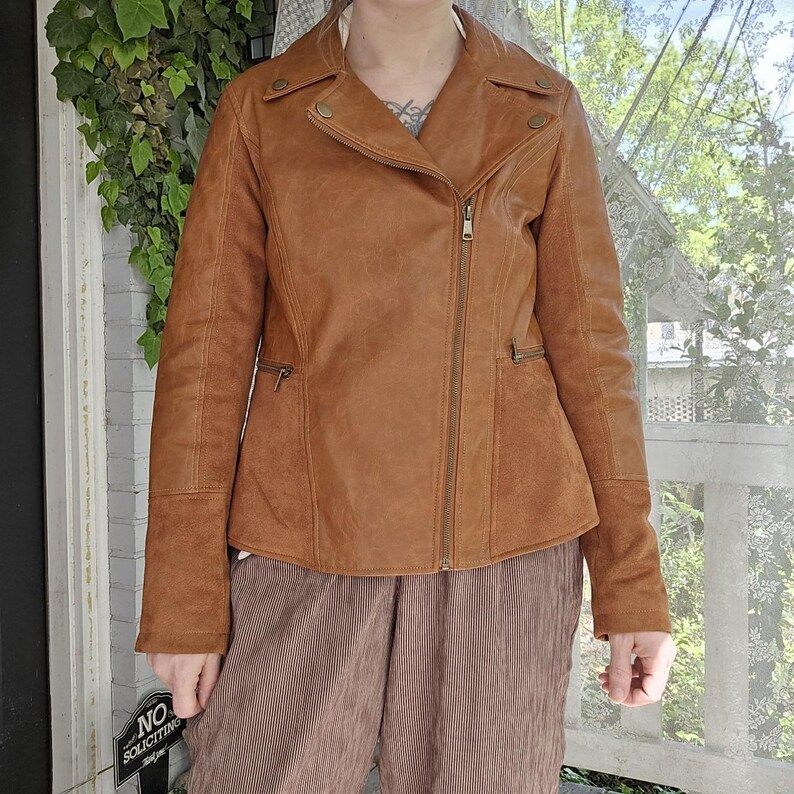 May include: A brown leather jacket with an asymmetrical zipper closure, two zippered pockets, and button details on the collar and shoulders. The jacket is a warm, earthy tone and has a classic biker style.