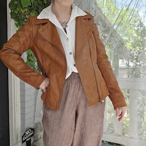 May include: A brown leather jacket with an asymmetrical zipper and a notched collar. The jacket has a classic biker style with a fitted silhouette and button details. The model is wearing a white collared shirt and brown corduroy pants.
