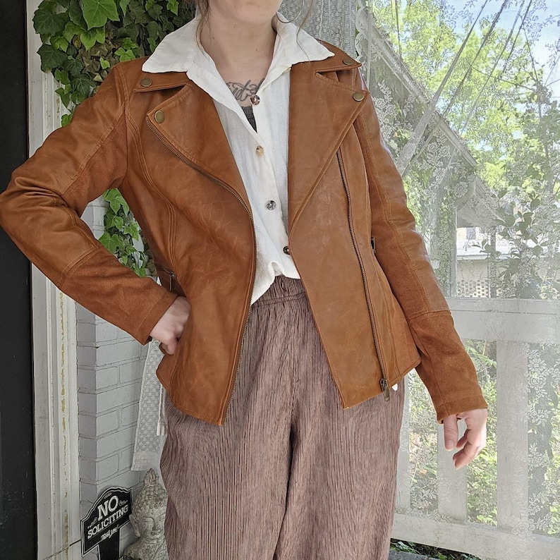 May include: A brown leather jacket with an asymmetrical zipper and a notched collar. The jacket has a classic biker style with a fitted silhouette and button details. The model is wearing a white collared shirt and brown corduroy pants.