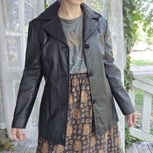 May include: A black leather blazer with a notched collar and three-button fastening. The blazer is worn over a grey graphic t-shirt and a patterned skirt with a brown and gold design. The outfit is set against a backdrop of a white curtain and a porch.