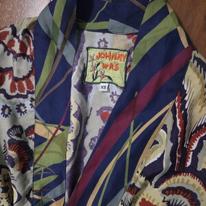 Pode incluir: Um roup&atilde;o da marca Johnny Was com uma estampa floral e bot&acirc;nica vibrante em tons de azul marinho, verde oliva, creme e vermelho escuro. A pe&ccedil;a possui uma gola larga e uma etiqueta no decote com o texto Johnny Was e uma etiqueta de tamanho XS.