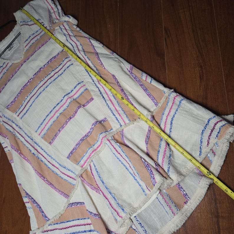 May include: A cream-colored dress with horizontal stripes in shades of peach, blue, and pink. The dress has a V-neck and a fringed hem. A yellow measuring tape is laid across the dress.