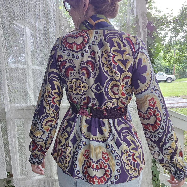 Pode incluir: Uma blusa de manga comprida com um padr&atilde;o repetitivo em tons de roxo, creme, vermelho e amarelo. A pe&ccedil;a inclui uma sec&ccedil;&atilde;o de cintura franzida e el&aacute;stica e uma gola alta, exibindo um design floral detalhado e intrincado.
