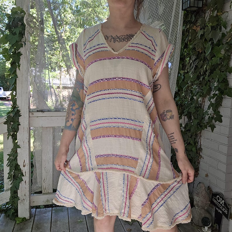 May include: A cream-colored, short-sleeved dress with horizontal stripes in peach, red, and blue. The dress has a V-neck and a ruffled hemline. The fabric appears to be lightweight and textured, suitable for warm weather.