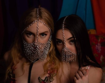 Puede incluir: Dos mujeres con máscaras faciales de malla plateada, una con un sujetador bordado con flores, la otra con una blusa rosa. Las mujeres posan juntas en una habitación con poca luz y tela colorida en el fondo.