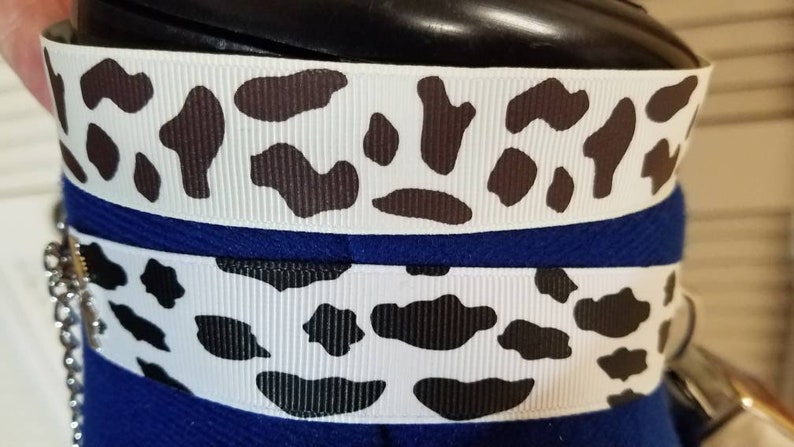May include: Two white ribbons with a black and white cow print design. The ribbons are layered on top of each other and are attached to a blue fabric.