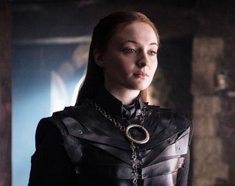 Puede incluir: Una mujer con el pelo largo y rojo que lleva un atuendo de cuero negro con un collar de cadena y anillo plateado.