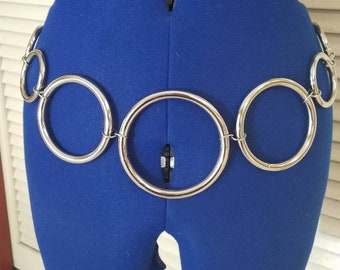 Puede incluir: Un cinturón de cadena plateado con múltiples eslabones grandes y redondos. El cinturón se lleva en un maniquí de tela azul.