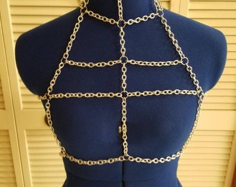Puede incluir: Joyería de cuerpo de cadena plateada con un diseño geométrico. La cadena está hecha de pequeños anillos unidos y se usa en un maniquí azul.
