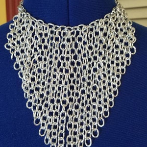 May include: A silver chainmail necklace with a V-shaped design. The necklace is made of many small, interconnected metal rings.