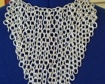 Puede incluir: Un collar de malla de cadena plateada con un diseño en forma de V. El collar está hecho de muchos anillos metálicos pequeños interconectados.
