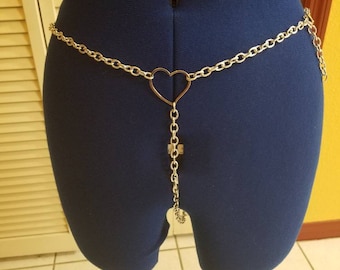 Puede incluir: Un cinturón de cadena plateado con un colgante en forma de corazón. La cadena está unida a un cierre en la parte posterior y el colgante cuelga hacia abajo en la parte delantera.