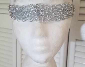 Puede incluir: Una diadema de cota de malla plateada se muestra sobre una cabeza de maniquí blanca. La diadema está hecha de pequeños anillos metálicos entrelazados, creando una banda ancha en la frente. El fondo es una puerta blanca con persianas.