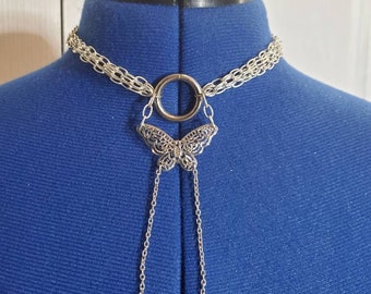 Puede incluir: Collar gargantilla de cadena plateada con un colgante de mariposa y un anillo grande. El collar está en un maniquí azul.