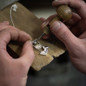 May include: A close-up of a person's hands working on a silver necklace with a geometric pendant. The person is using a small tool to attach the pendant to the chain.