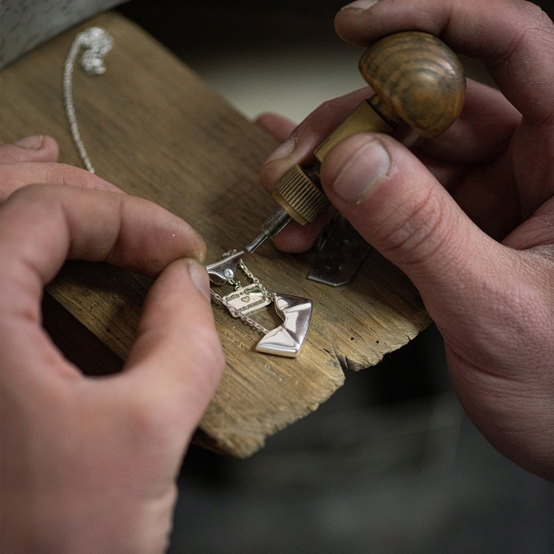 May include: A close-up of a person's hands working on a silver necklace with a geometric pendant. The person is using a small tool to attach the pendant to the chain.