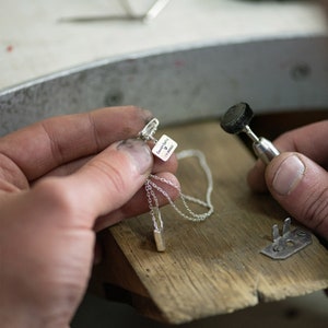 May include: A silver necklace with a small rectangular pendant that says "David & Carla 1.1.2023" with a heart symbol. The necklace is being held by a person's hand, and a small tool with a black sanding wheel is in the background.