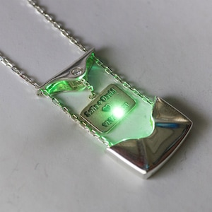 May include: A silver necklace with a green pendant that has a small diamond and a charm with the words "Sofia + David" and a heart. The charm is glowing green.
