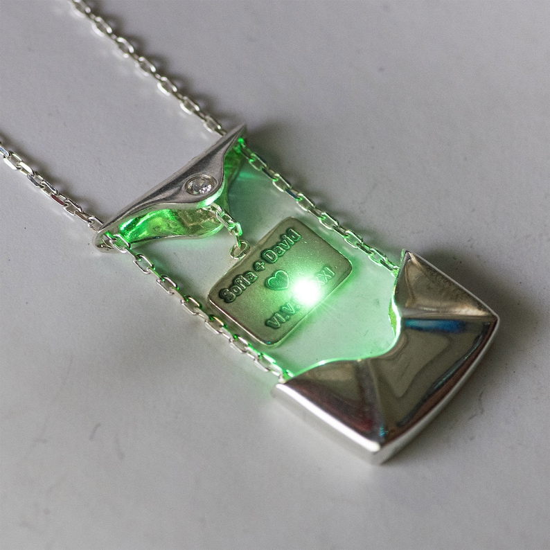 May include: A silver necklace with a green pendant that has a small diamond and a charm with the words "Sofia + David" and a heart. The charm is glowing green.