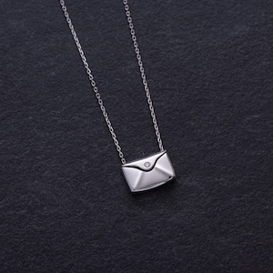 May include: A silver necklace with a small envelope pendant featuring a single diamond. The necklace is on a black background.