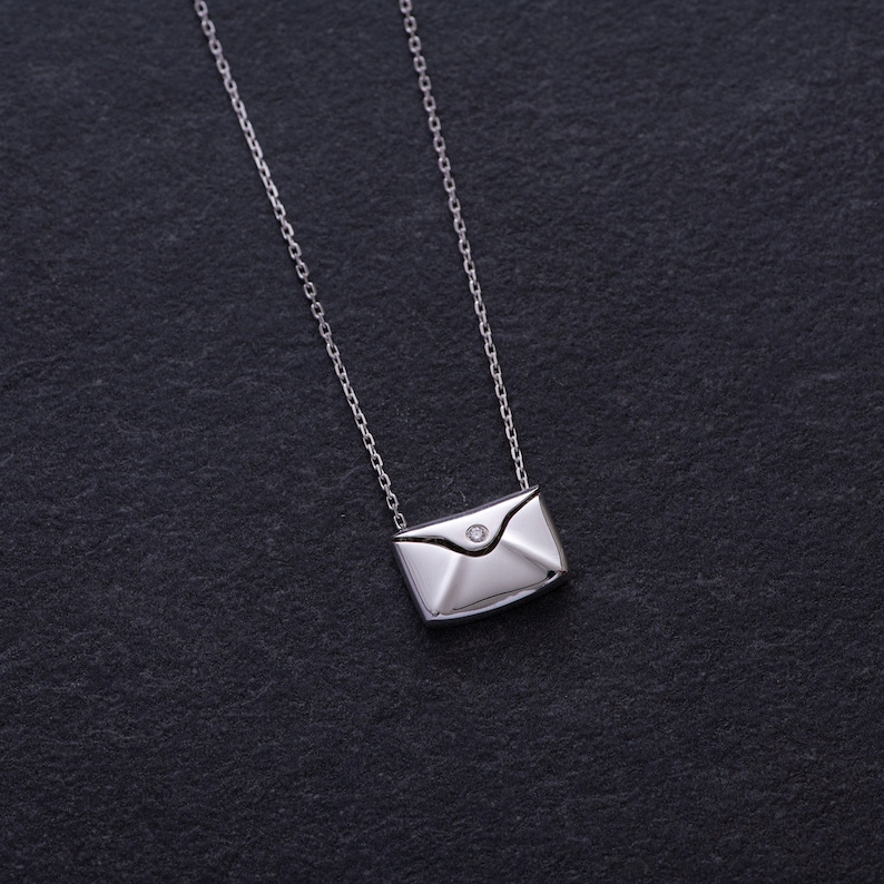 May include: A silver necklace with a small envelope pendant featuring a single diamond. The necklace is on a black background.
