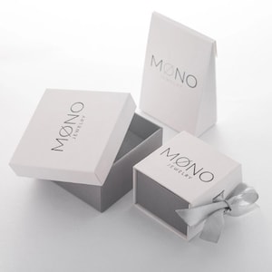 May include: Three white boxes with the logo "MONO JEWELRY" printed on them. Two of the boxes are square and one is a triangular box. The square boxes have a gray interior and the triangular box is white inside. One of the square boxes has a silver ribbon tied around it.