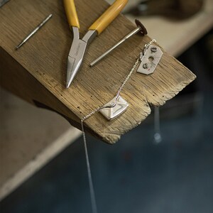 May include: A silver necklace with a small envelope pendant featuring a single diamond. The necklace is lying on a wooden surface with a pair of pliers and a small metal tool.