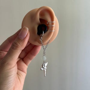 May include: A silicone ear plug with a dangling silver-toned chain attachment featuring a small, translucent white bead and a silver-toned fairy charm. The ear model also displays three thin, stacked silver-toned ear cuffs on the upper cartilage.