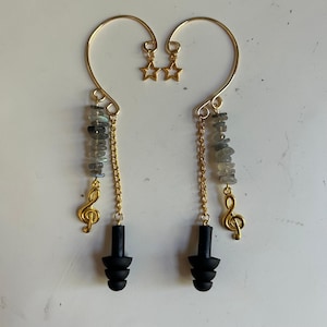 May include: A pair of gold-toned ear cuffs featuring a curved wire frame with a small star charm. Each cuff includes a dangling gold chain, a strand of grey stone chips, a gold treble clef charm, and a black three-tiered silicone earplug.