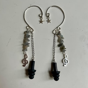Puede incluir: Un par de pendientes tipo ear cuff plateados con marco de alambre curvo y una pequeña estrella. Cada uno incluye una cadena con un tapón auditivo negro de tres bridas, cuentas de labradorita gris y un dije de clave de sol plateado.