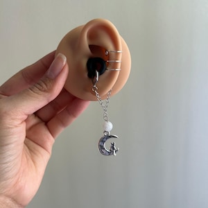 May include: A silver-toned ear cuff with three bands, a black ear plug, and a dangling chain attachment featuring a white bead and a silver crescent moon charm with a small cat silhouette sitting on the edge of the moon, displayed on an ear model.
